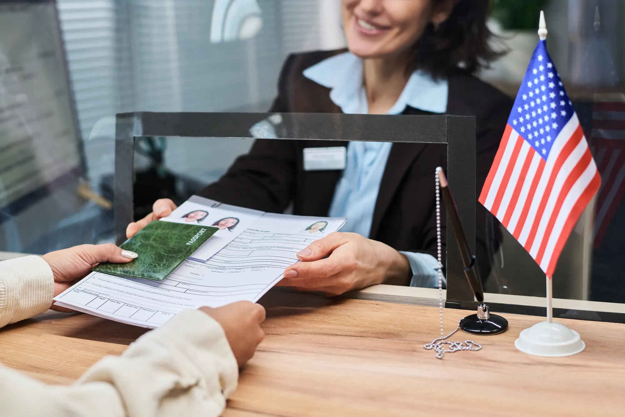 Manos entregando un pasaporte y formularios migratorios en una ventanilla, junto a una bandera de Estados Unidos